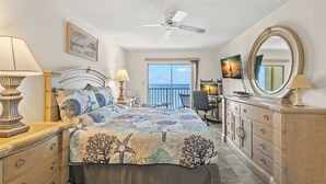 Un dormitorio con cama, lámparas de noche, ventilador de techo, cómoda, escritorio y un gran ventanal con vistas al océano.