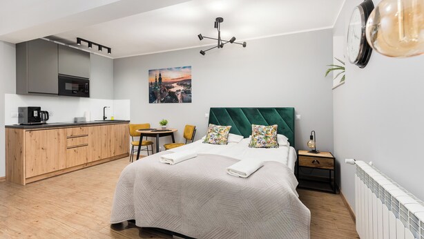 A modern hotel room with a bed, a desk, a kitchenette, and a view of a cityscape. - Underground Basilica Apartment (Kraków)