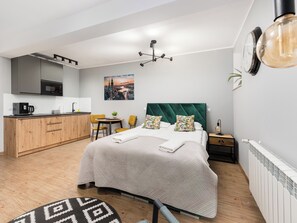 A modern hotel room with a bed, a desk, a kitchenette, and a view of a cityscape.