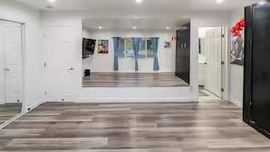 A spacious room with a wooden floor, white walls, and a large mirror. There are two doors, one on each side, and a window with blue curtains. - The Shalimar Casita, Fits 6 Guests, Smart Tvs, Remodeled Unit, Full Size Kitchen (Riverside)