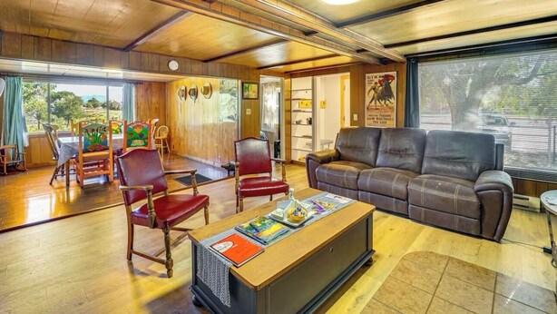 Una sala de estar acogedora con un sofá de cuero marrón, sillas de madera, una mesa de centro con libros y un gran ventanal con vistas al exterior.