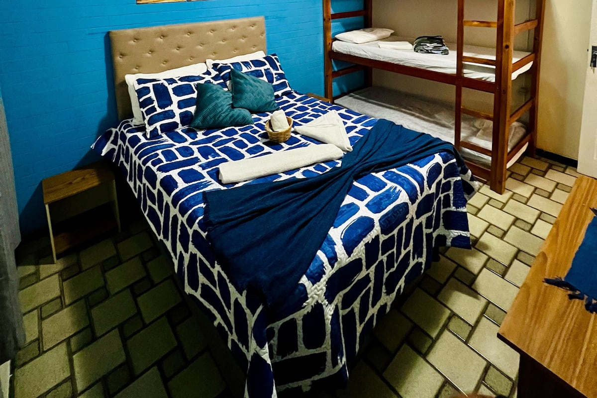 A bedroom with a bed, a bunk bed, a nightstand, a wall with a sunset painting, and a tiled floor.