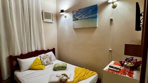 Une chambre d’hôtel avec un plafond en bois, un lit recouvert d’une courtepointe jaune et blanche, un tableau représentant une plage et une petite table sur laquelle se trouvent une télécommande et des en-cas.