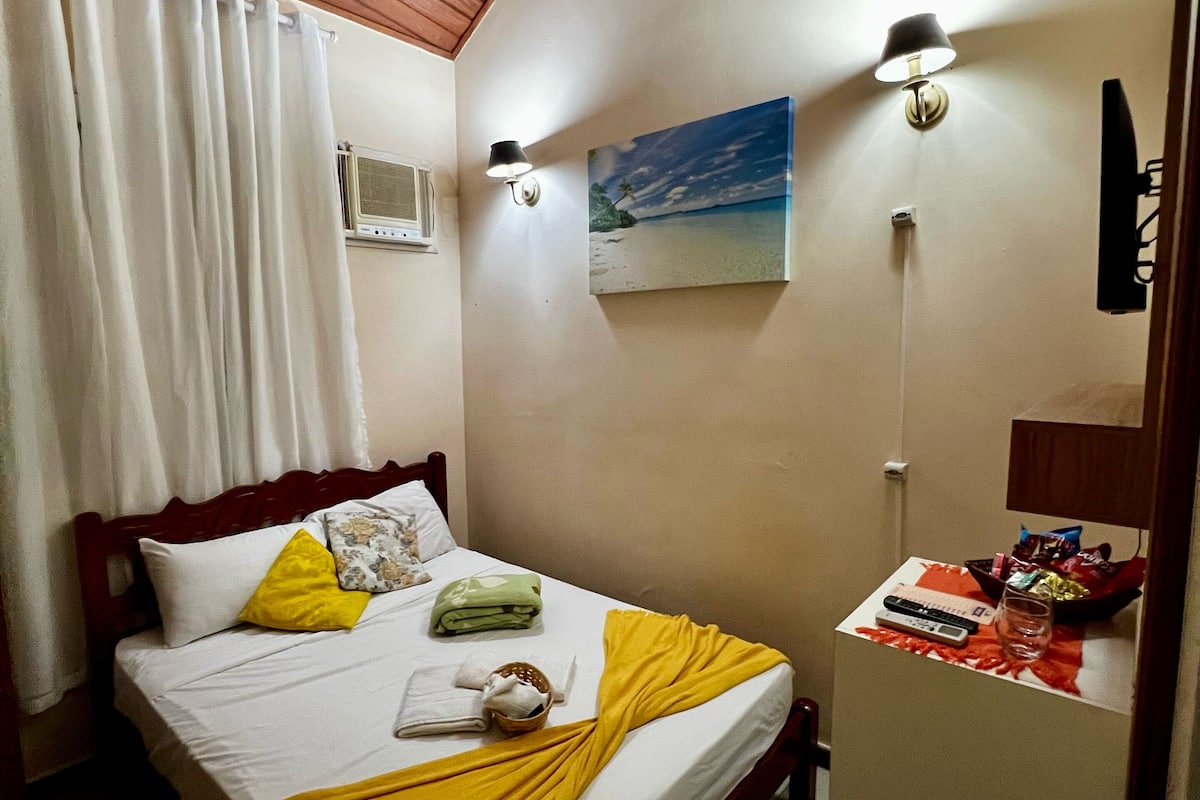 A hotel room with a wooden ceiling, a bed with a yellow and white bedspread, a painting of a beach, and a small table with a remote control and snacks.