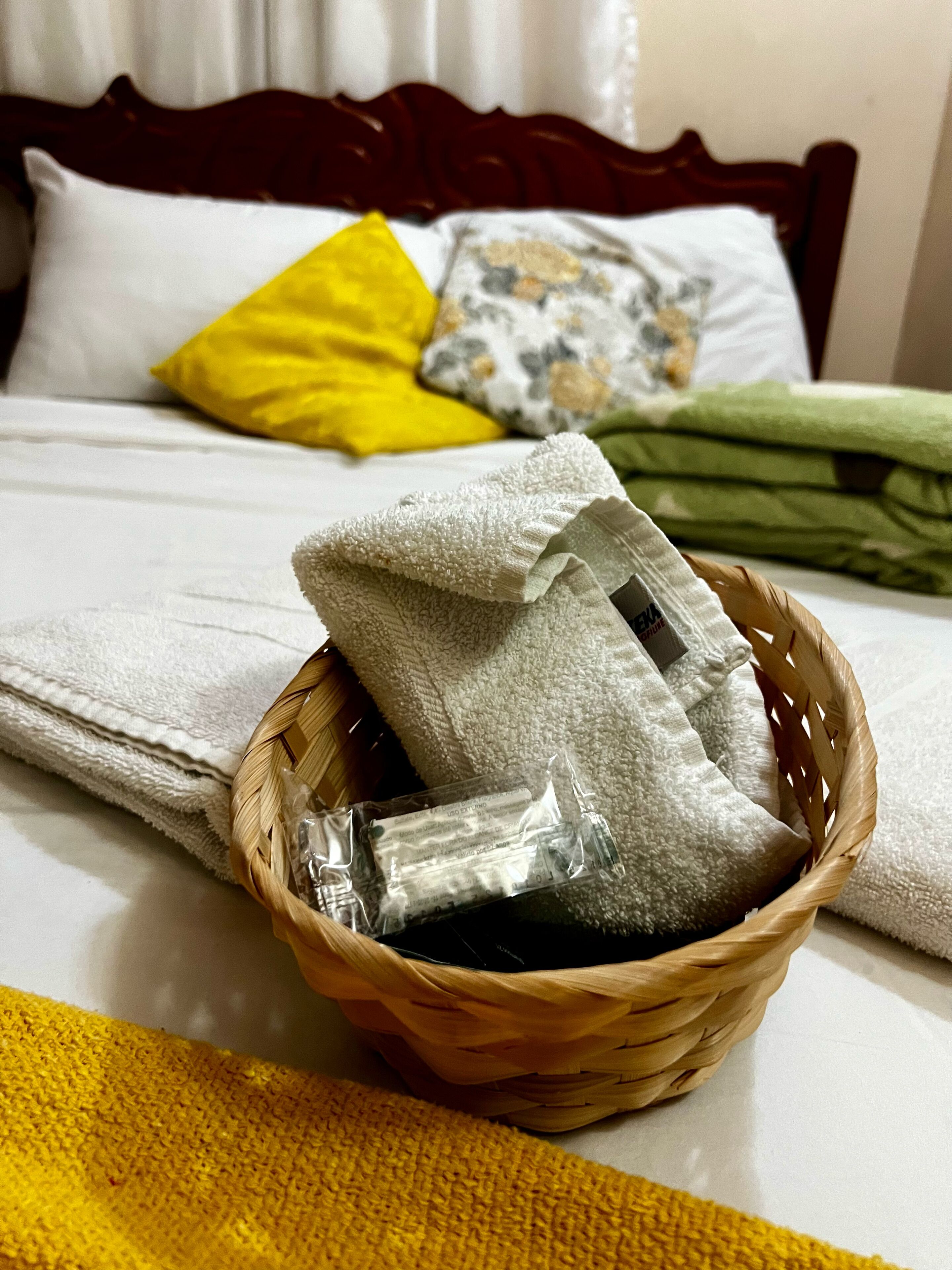 A bed with white and yellow pillows, a green blanket, and a basket containing folded towels and a black object.