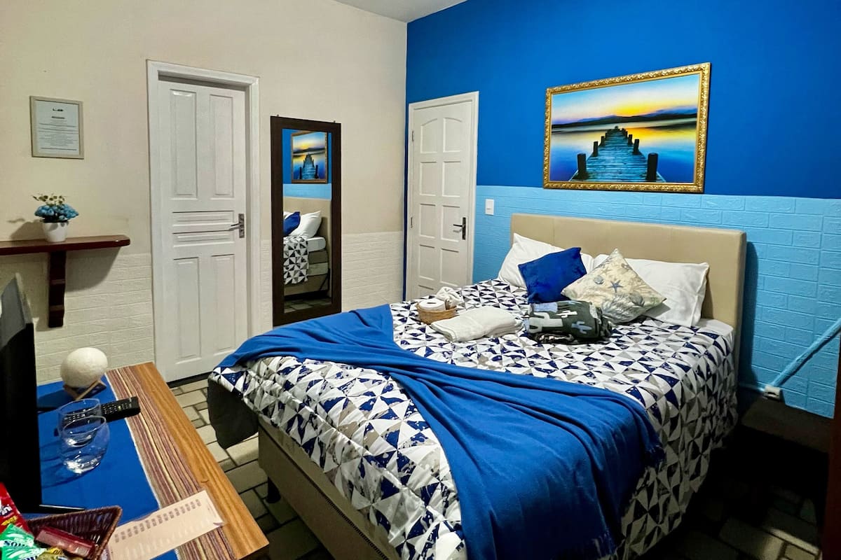 A bedroom with a bed, a desk, a chair, a television, and a painting on the wall.