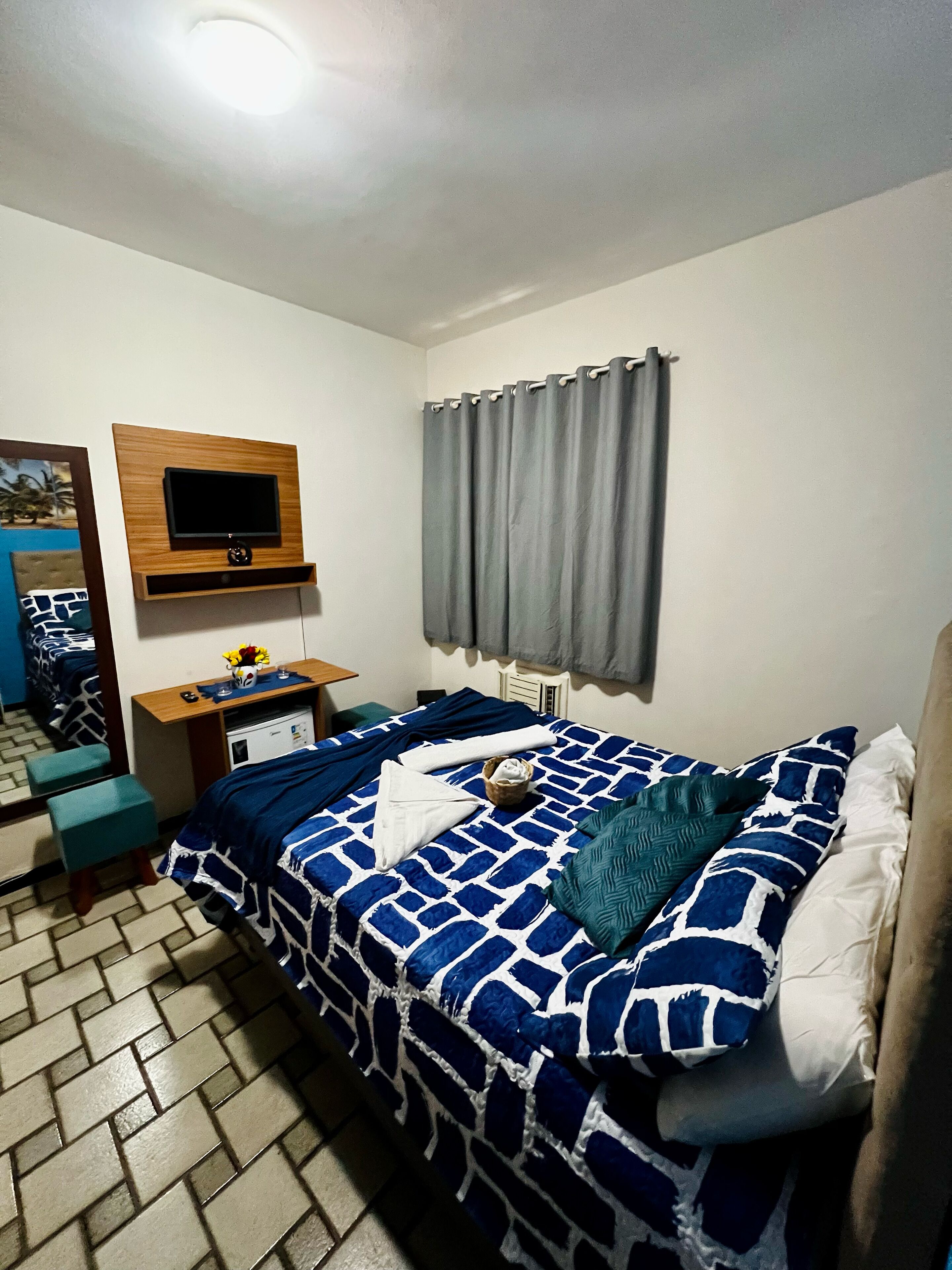 A neatly arranged bedroom with a bed, a bedside table, a mirror, a TV mounted on the wall, and a window with curtains.