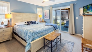 Een slaapkamer met een bed, nachtkastje, lamp, plafondventilator, raam met gordijnen, schilderij, stoel en uitzicht op een balkon.