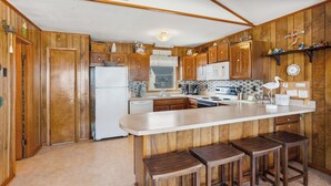 A kitchen with wooden cabinets, a white refrigerator, a large island with stools, and a ceiling fan. - 7050 Sailor's Rest at Nags Head * Oceanfront (Nags Head)