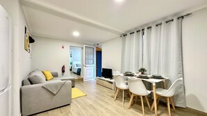A modern living room with a grey sofa, a dining table with chairs, and a TV. - Casa do Perestrelo - Guest House (Machico)