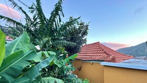 A yellow building with a red-tiled roof, surrounded by lush green foliage and a mountainous backdrop. - Casa do Perestrelo - Guest House (Machico)