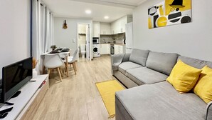 A modern apartment with a grey sofa, yellow pillows, a dining area, a kitchen, and a TV. - Casa do Perestrelo - Guest House (Machico)