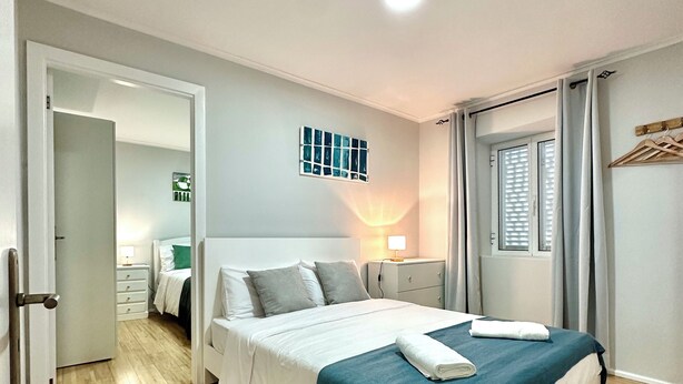 A modern bedroom with a large bed, a bedside table, a wall-mounted light, and a view of a hallway. - Casa do Perestrelo - Guest House (Machico)