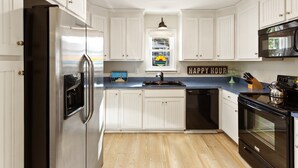 A modern kitchen with white cabinetry, stainless steel appliances, a black stove, and a window with a colorful view. - Home Sweet Helga: Beach & Sound Access (Kill Devil Hills)