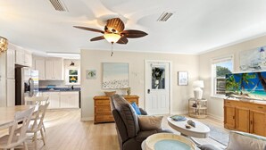 A spacious living area with a large sofa, a dining table with chairs, a kitchen with white cabinets, and a ceiling fan. - Home Sweet Helga: Beach & Sound Access (Kill Devil Hills)
