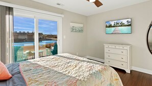 A bedroom with a bed, a dresser, a ceiling fan, a TV, and a view of a pool and beach.