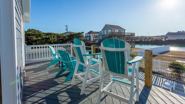A wooden deck with turquoise Adirondack chairs, a white railing, and a view of a house and water body.