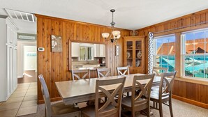 A dining area with a wooden table and chairs, a kitchen in the background, and a view of a beach through large windows.