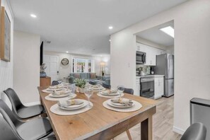 Un comedor con una mesa de madera dispuesta para cuatro, una cocina al fondo y una sala de estar visible a través de una puerta abierta.