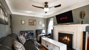 A living room with a fireplace, a black leather sofa, a white coffee table, a television mounted above the fireplace, and a ceiling fan. - Peaceful Pretty Art and Cinema Reclining Couches (Ferndale)