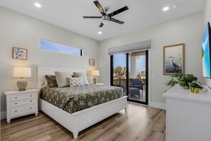 Une chambre à coucher comprenant un lit, des tables de chevet, un ventilateur de plafond, une fenêtre et une porte-fenêtre coulissante donnant sur un balcon.