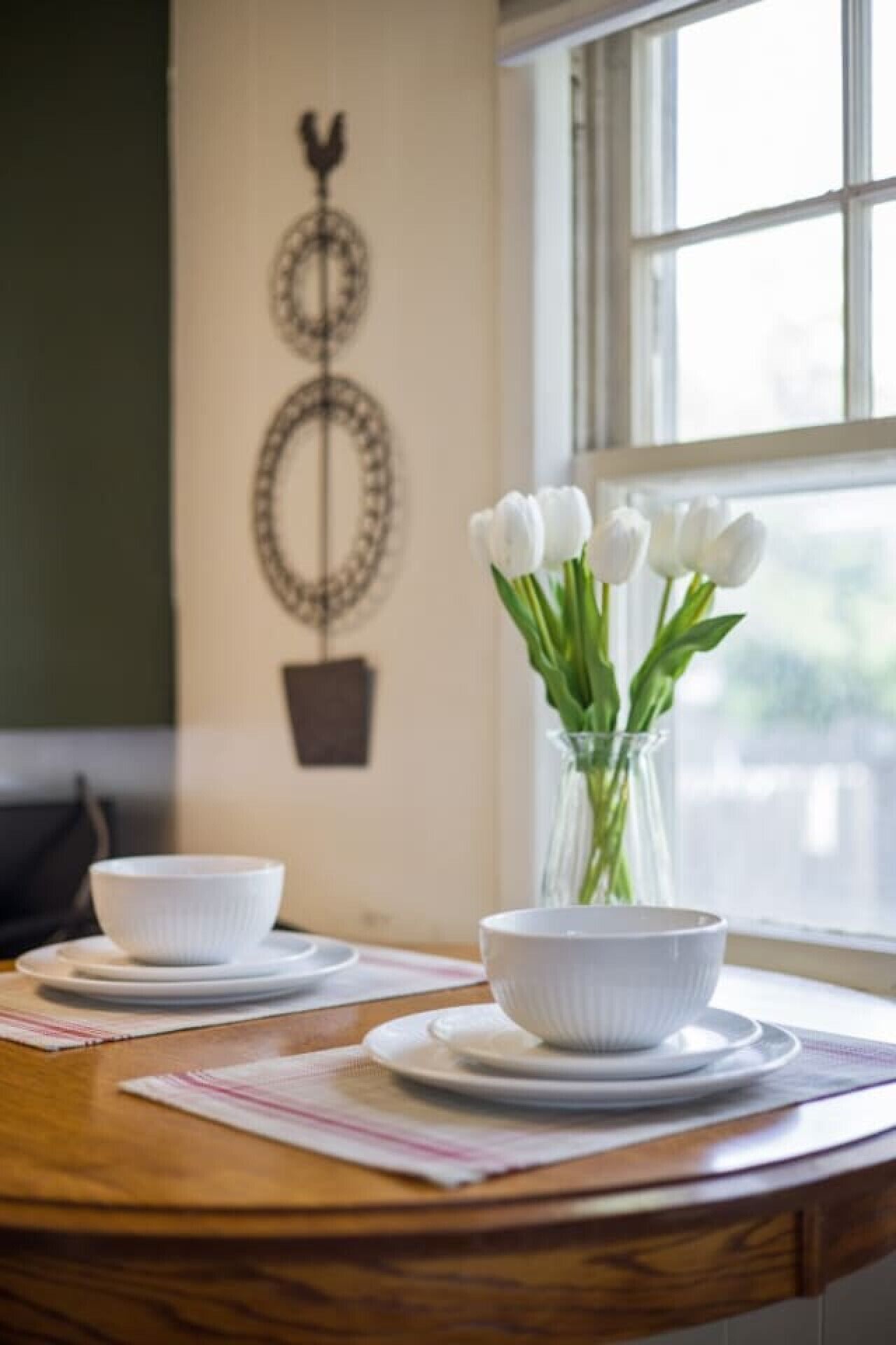 A dining table with two white bowls on plates, a vase with white tulips, and a wall-mounted decorative piece.