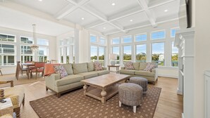 Un vaste salon comprenant un grand canapé d’angle, une table basse et des poufs. On y trouve également un coin repas avec une table et des chaises, ainsi que de grandes fenêtres offrant une vue sur l’extérieur.