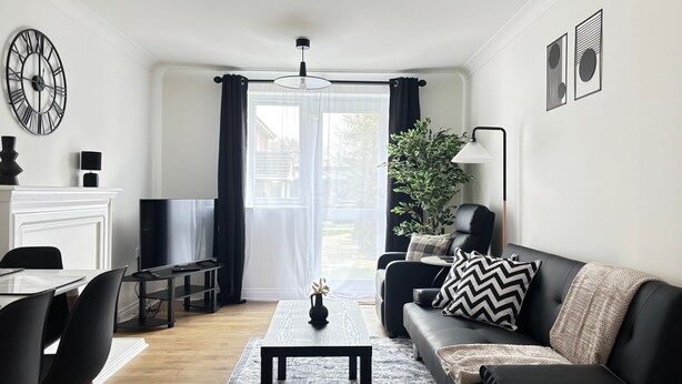 Un salon moderne avec un canapé noir, une table basse, un coin repas avec une table et des chaises, une cheminée et une grande fenêtre avec des rideaux.