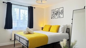 Une chambre à coucher comprenant un lit, un radiateur, une fenêtre avec des rideaux, un luminaire fixé au mur et des œuvres d’art encadrées.