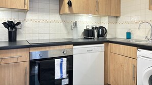 Une cuisine avec des meubles de rangement en bois, un plan de travail noir, un lave-vaisselle, une machine à laver et une machine à café.