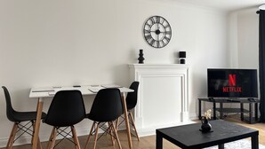 Un salon moderne avec une cheminée blanche, une horloge noire, une table à manger avec des chaises et un téléviseur affichant le logo de Netflix.