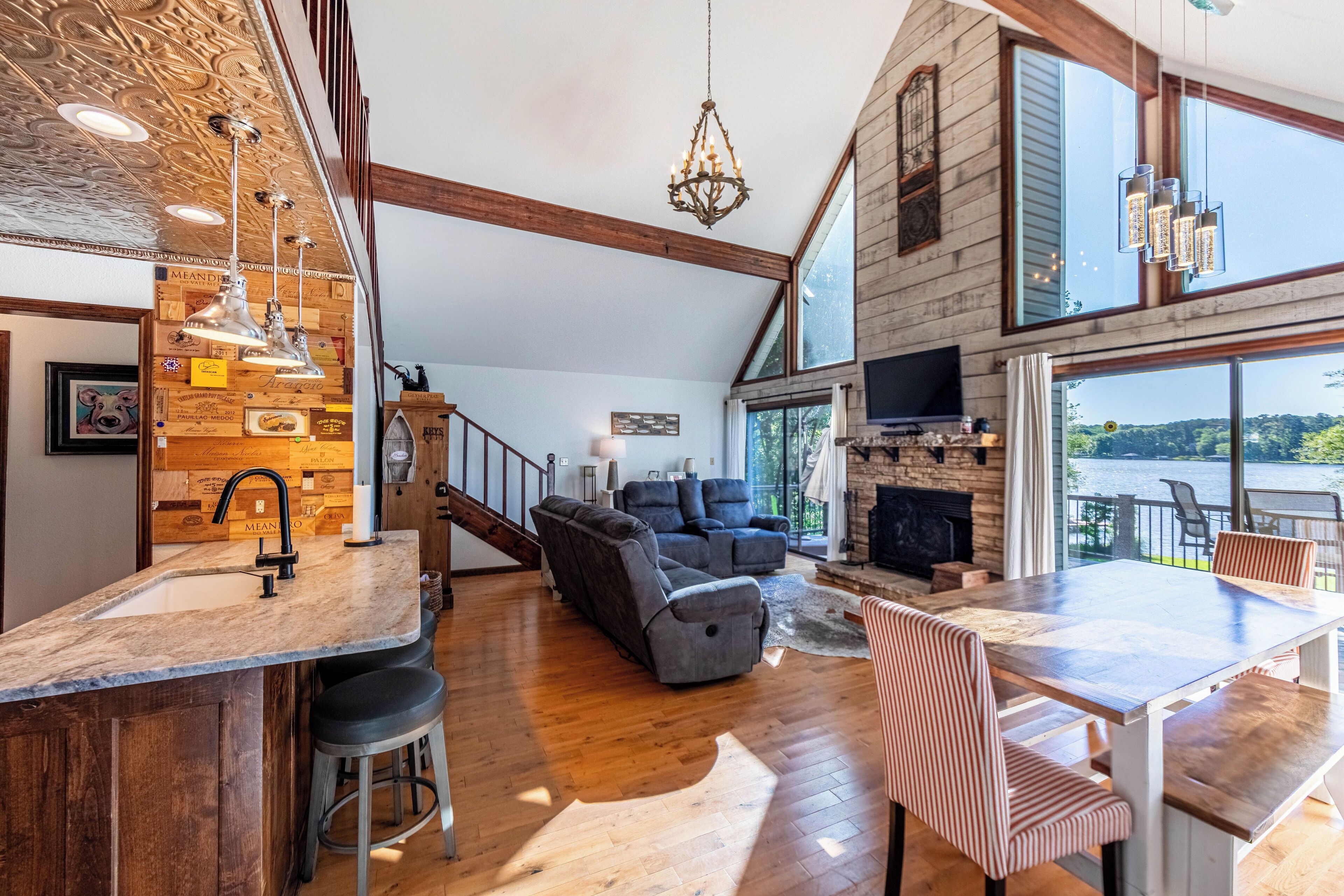 A spacious living area with a fireplace, a large window overlooking a body of water, and a modern kitchen with a bar.
