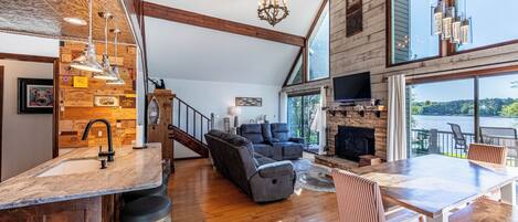 A spacious living area with a fireplace, a large window overlooking a body of water, and a modern kitchen with a bar.