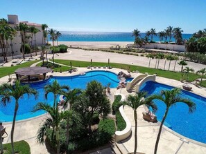 Un resort con una amplia piscina central, palmeras y vista al mar.