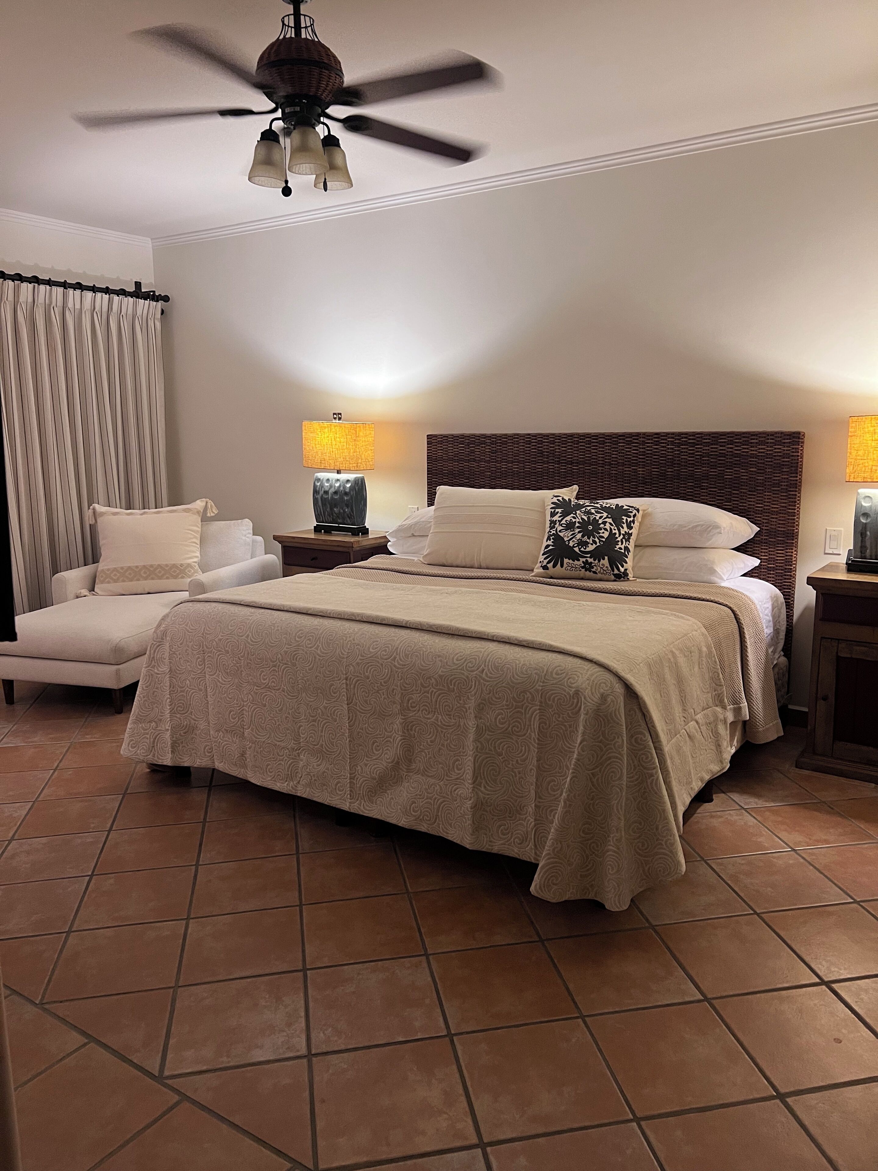 Un dormitorio con una cama grande, dos mesitas de noche con lámparas, un ventilador de techo y una silla.