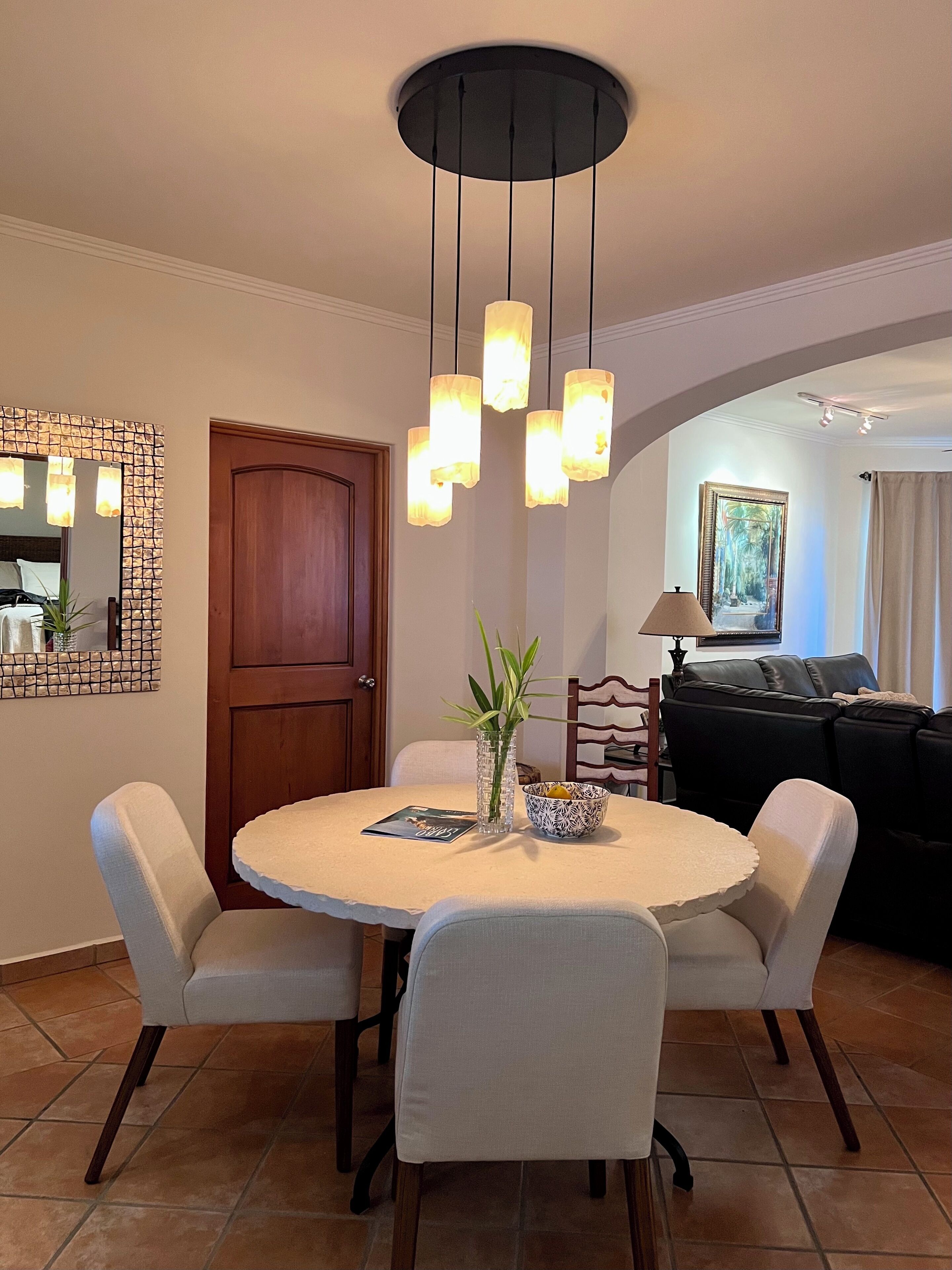 A dining area with a round table, four chairs, a vase with a plant, and a bowl of fruit. There is a wooden door and a black sofa in the background.