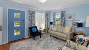 Una sala de estar con una puerta azul, un sillón negro con cojín azul, un sofá color crema y una alfombra estampada.