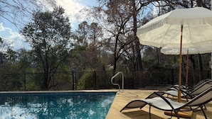 A swimming pool with lounge chairs and a white umbrella, surrounded by trees and a fence. - Creekside Escape – Spacious 5BR with Pool & BBQ (Columbus)