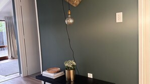 Une pièce avec un mur vert foncé, un plancher en bois, une table noire ornée d’une plante, un panier en osier et un luminaire fixé au mur.
