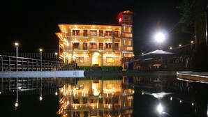 A brightly lit building with a reflection in the water, featuring multiple balconies and a prominent tower. - SUKOON DK (Jakholi)