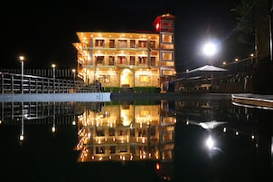 Um edifício bem iluminado com reflexo na água, apresentando várias varandas e uma torre imponente.