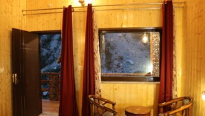 A wooden cabin interior with a large window, two wooden chairs, and a door leading outside. - SUKOON DK (Jakholi)