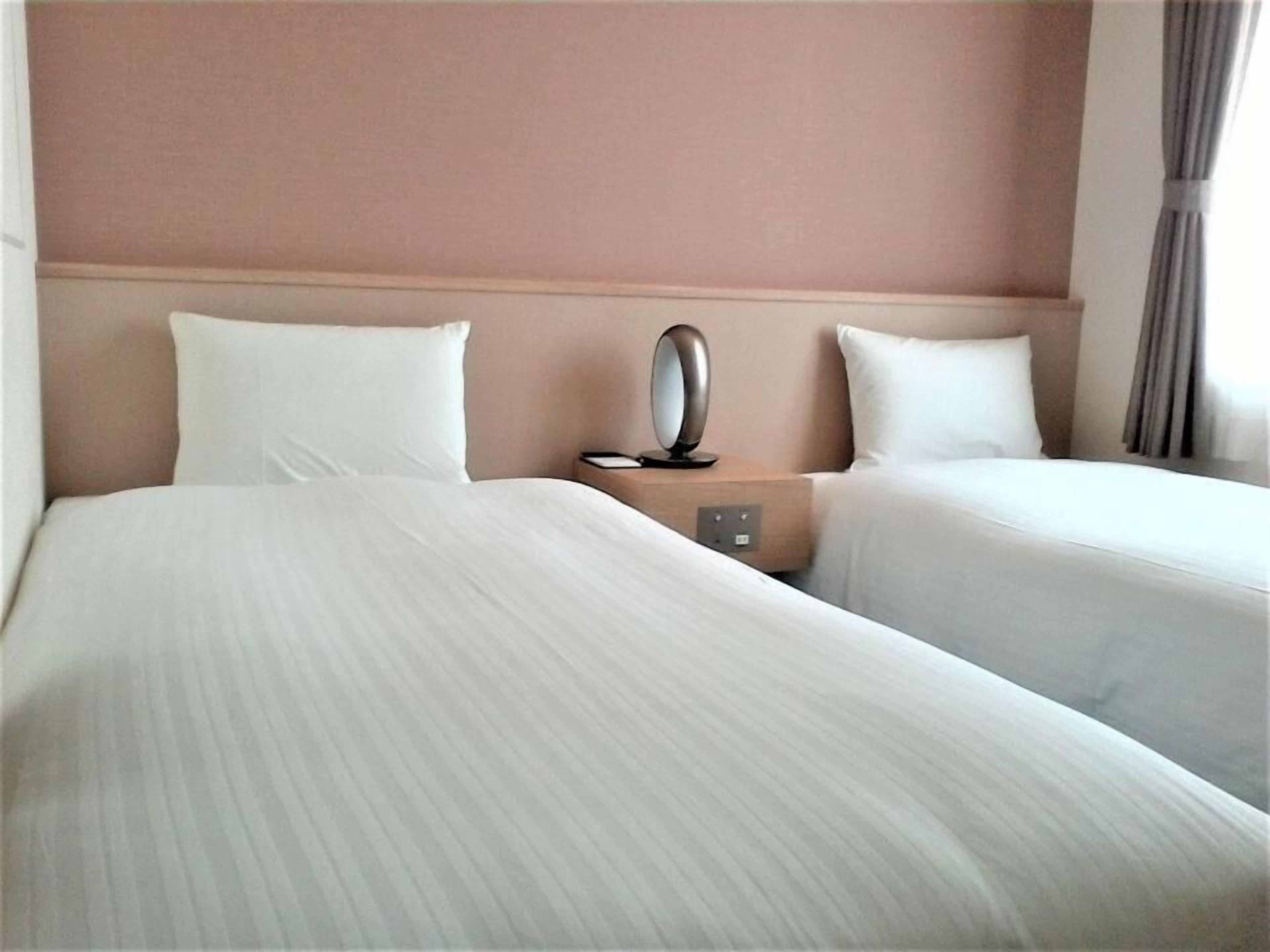 Quarto de hotel com duas camas, uma mesa de cabeceira com espelho e uma parede lisa.