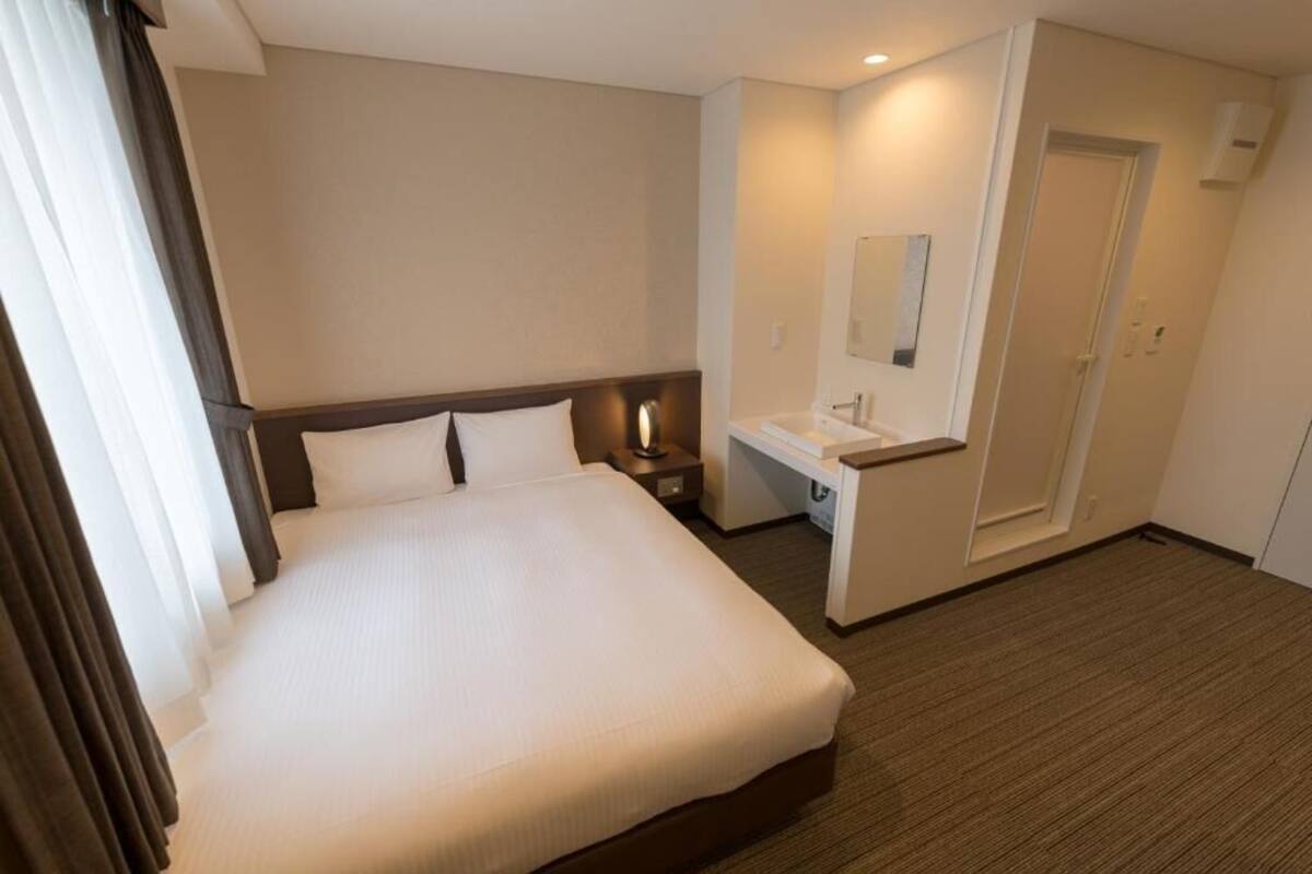 Une chambre d’hôtel avec un grand lit, une table de chevet, un petit bureau avec une chaise, un miroir et une porte.