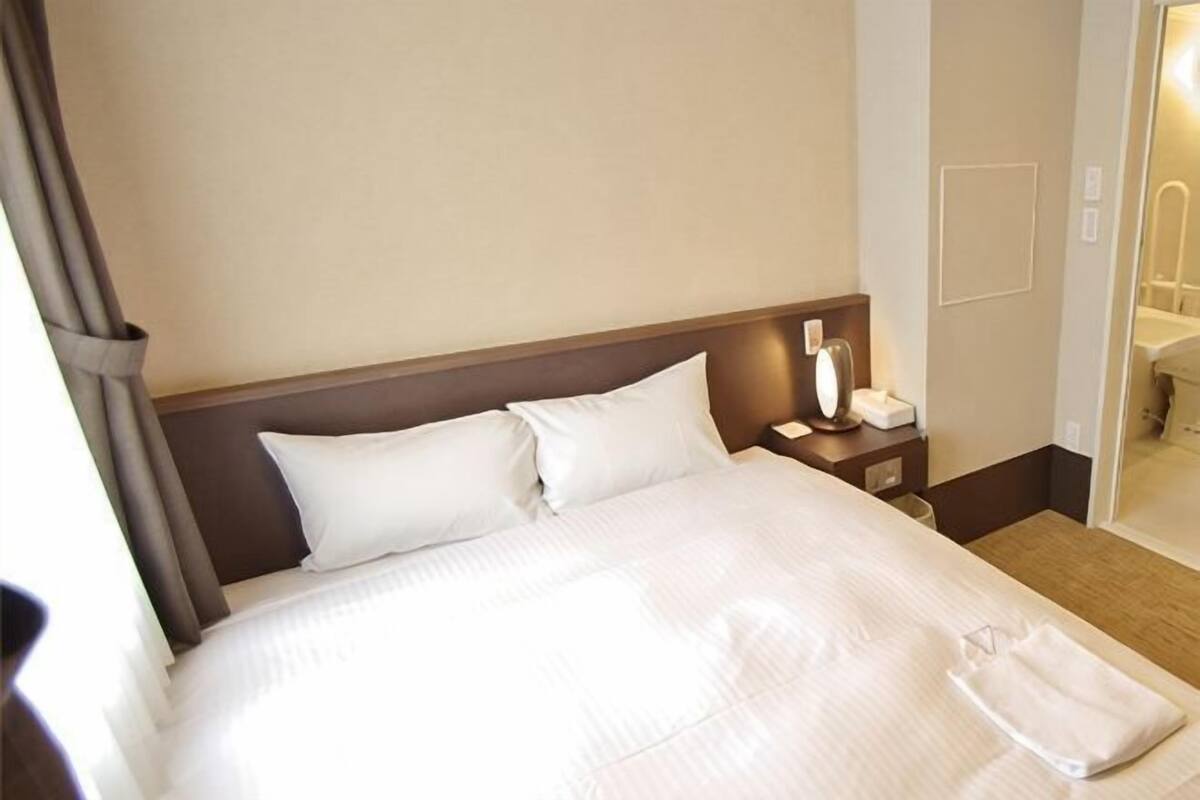 Une chambre d’hôtel avec un grand lit, une table de chevet avec une lampe et une salle de bain visible à travers une porte ouverte.