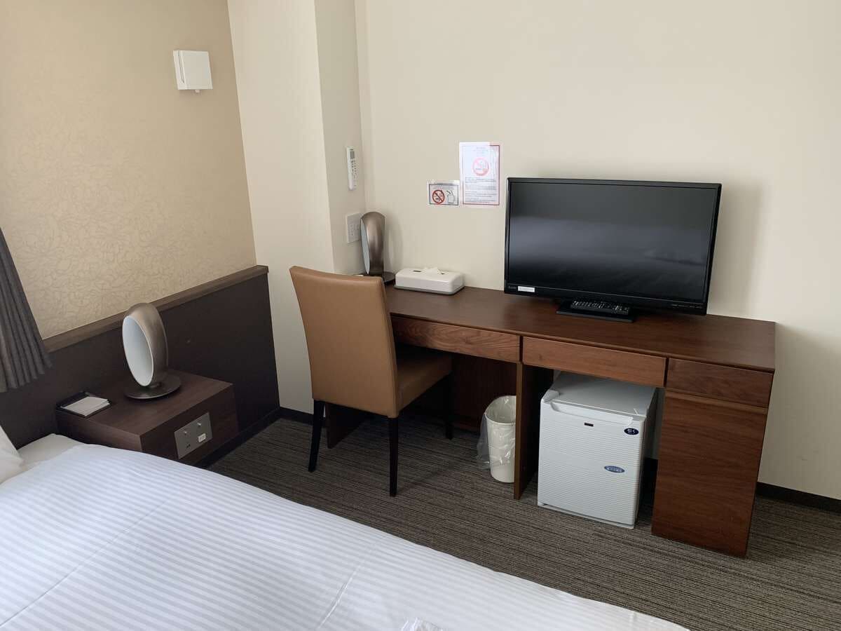 A hotel room with a bed, a desk with a television, a chair, a small table with a lamp, and a trash bin.