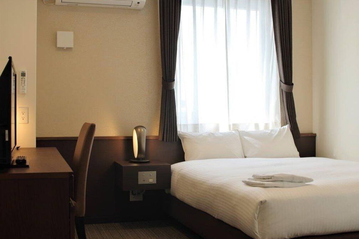 Une chambre d’hôtel avec un lit, un bureau, une chaise, une lampe et une fenêtre avec des rideaux.