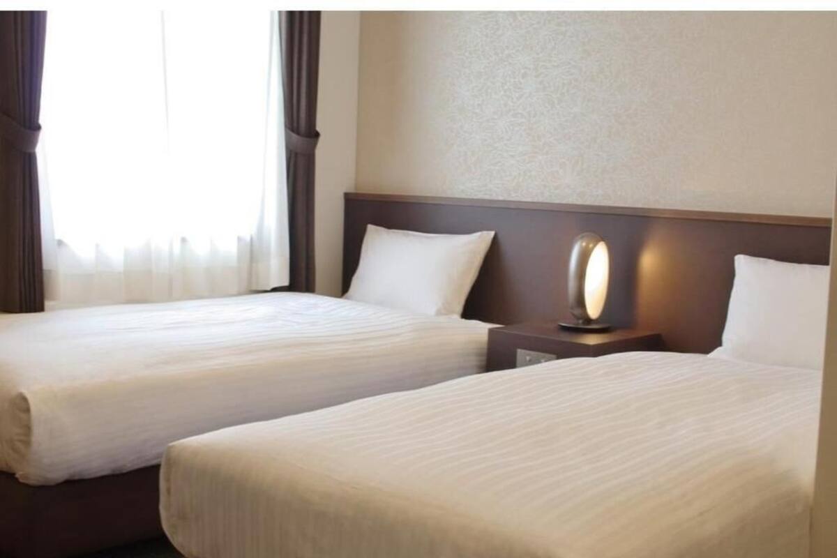 Une chambre d’hôtel avec deux lits, une table de chevet avec une lampe et une fenêtre avec des rideaux.
