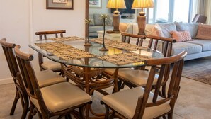 A dining area with a glass table surrounded by wooden chairs, a chandelier, and a sofa in the background. - Coquina B101 Ground-Floor Coastal Condo (Saint Augustine)