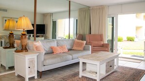 A living room with a sofa, armchairs, a coffee table, a TV, and a large mirror. - Coquina B101 Ground-Floor Coastal Condo (Saint Augustine)
